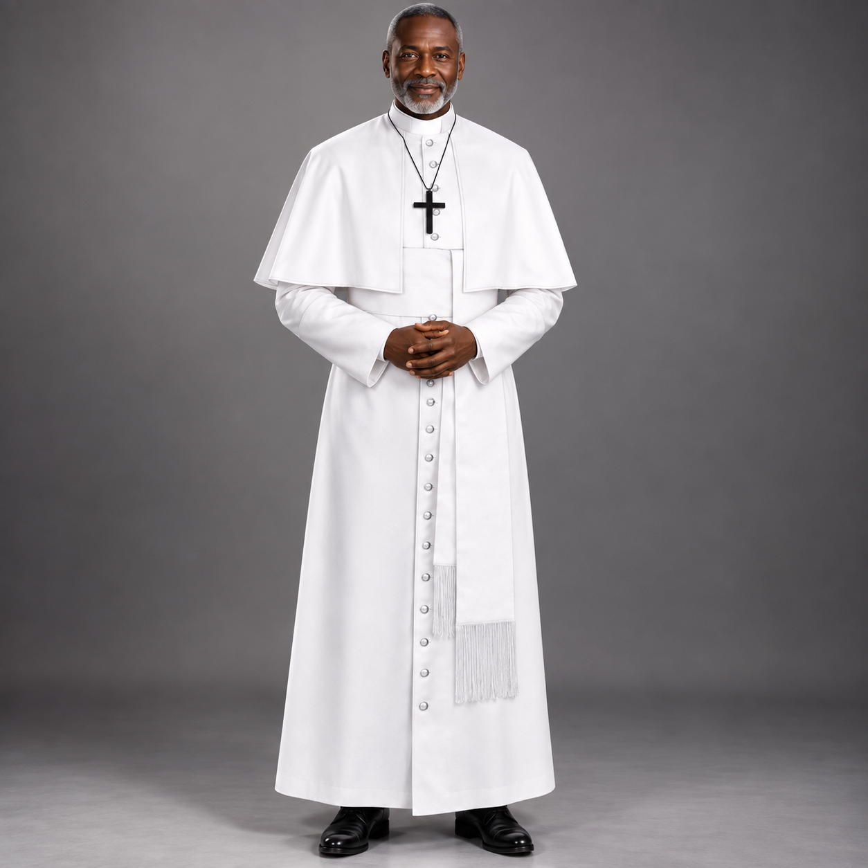 A man wearing a white priestly robe with a black cross necklace, standing against a solid gray studio background.