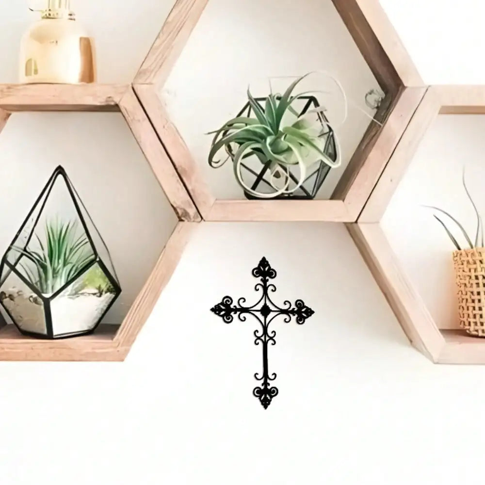 A black metal wall cross with ornate scrollwork hangs on a white wall below a hexagonal wooden shelf. The shelf holds small potted plants, including an air plant in a geometric glass terrarium and another in a woven pot.