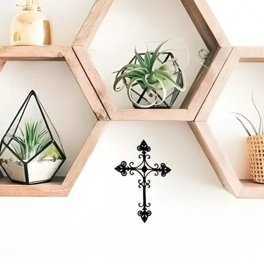 A black metal wall cross with ornate scrollwork hangs on a white wall below a hexagonal wooden shelf. The shelf holds small potted plants, including an air plant in a geometric glass terrarium and another in a woven pot.
