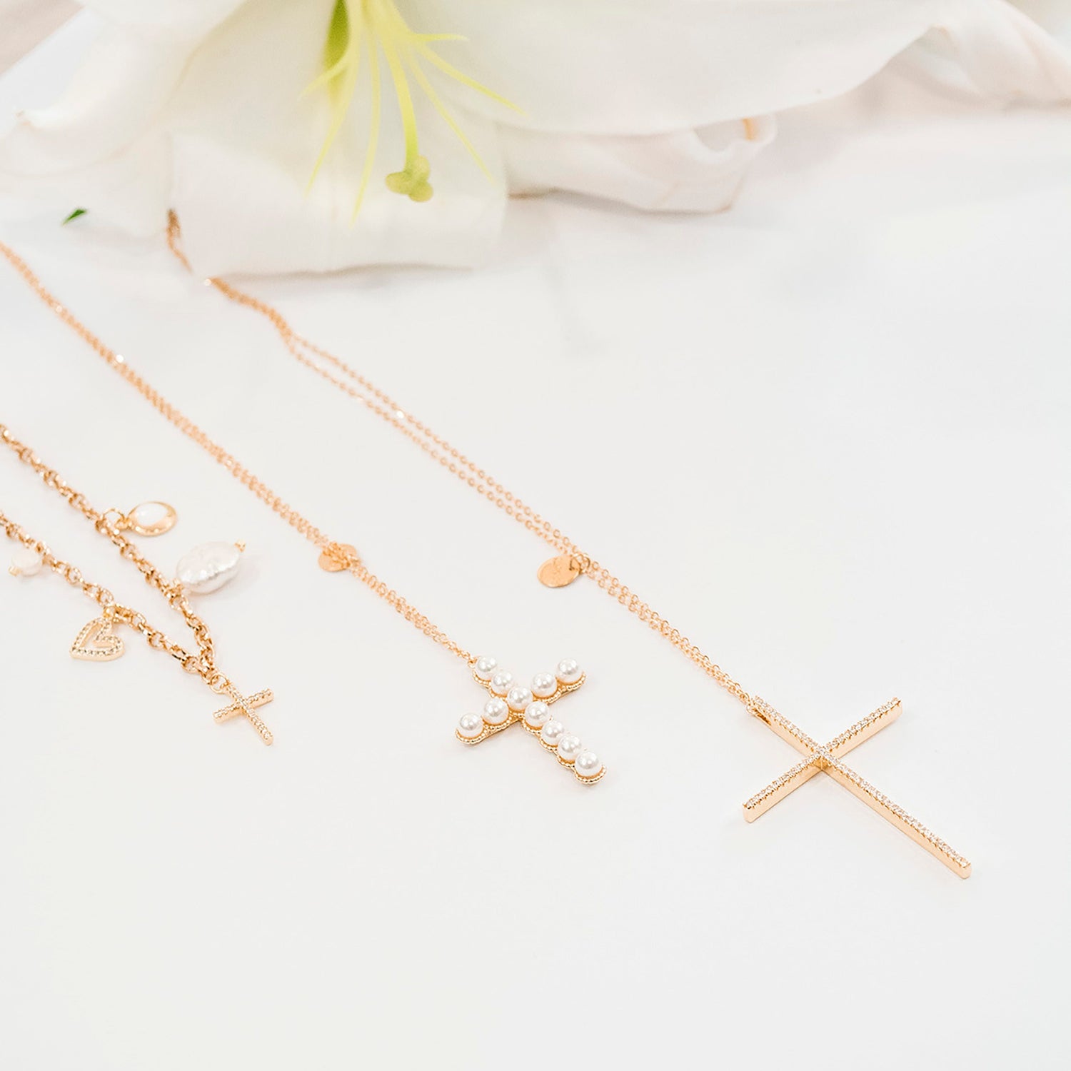 Three delicate gold-toned necklaces with cross and heart charms are arranged on a white surface with white lilies in the background.
